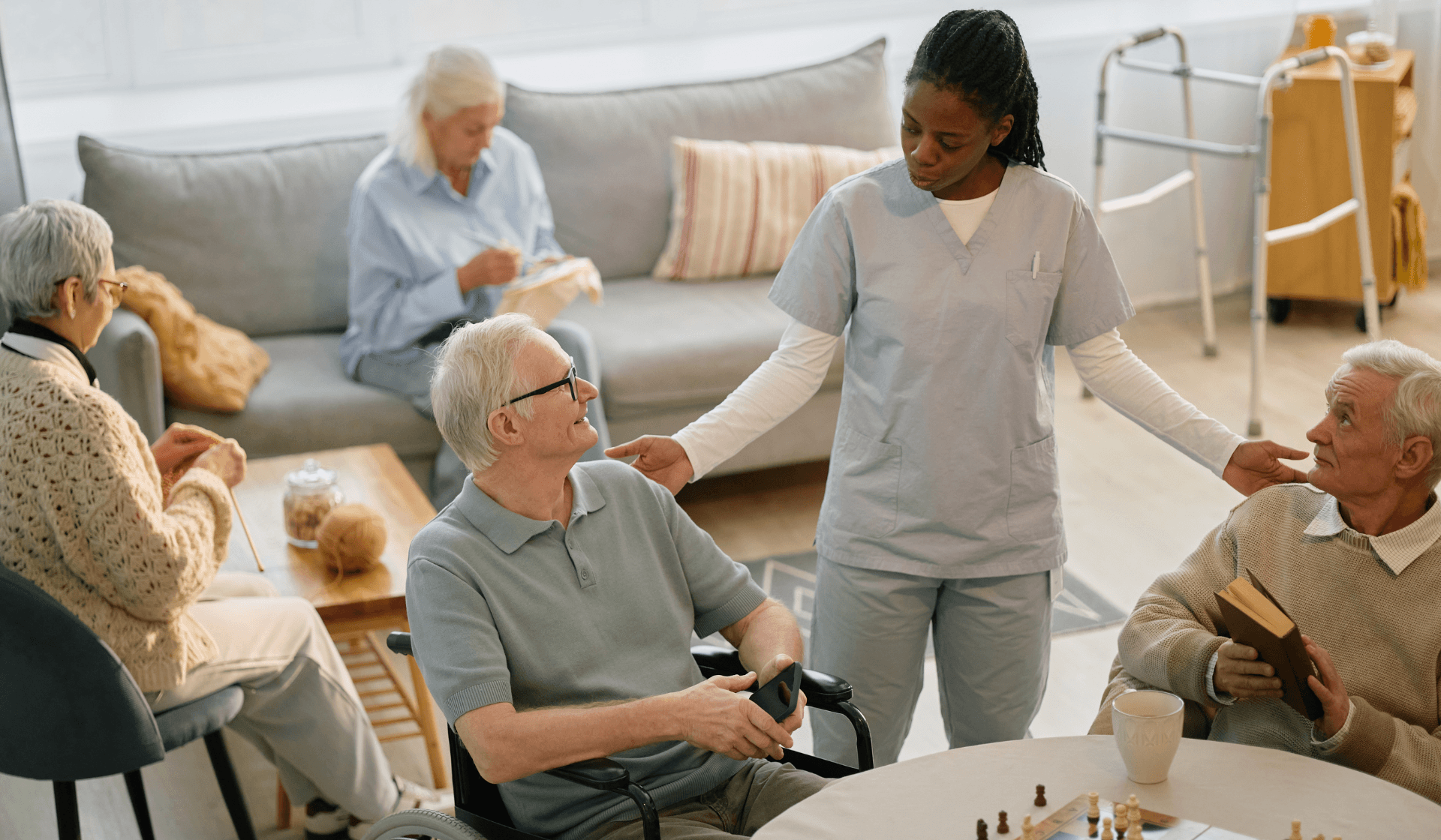 senior aid talking to two senior men at an assisted living home, with two senior women behind them knitting