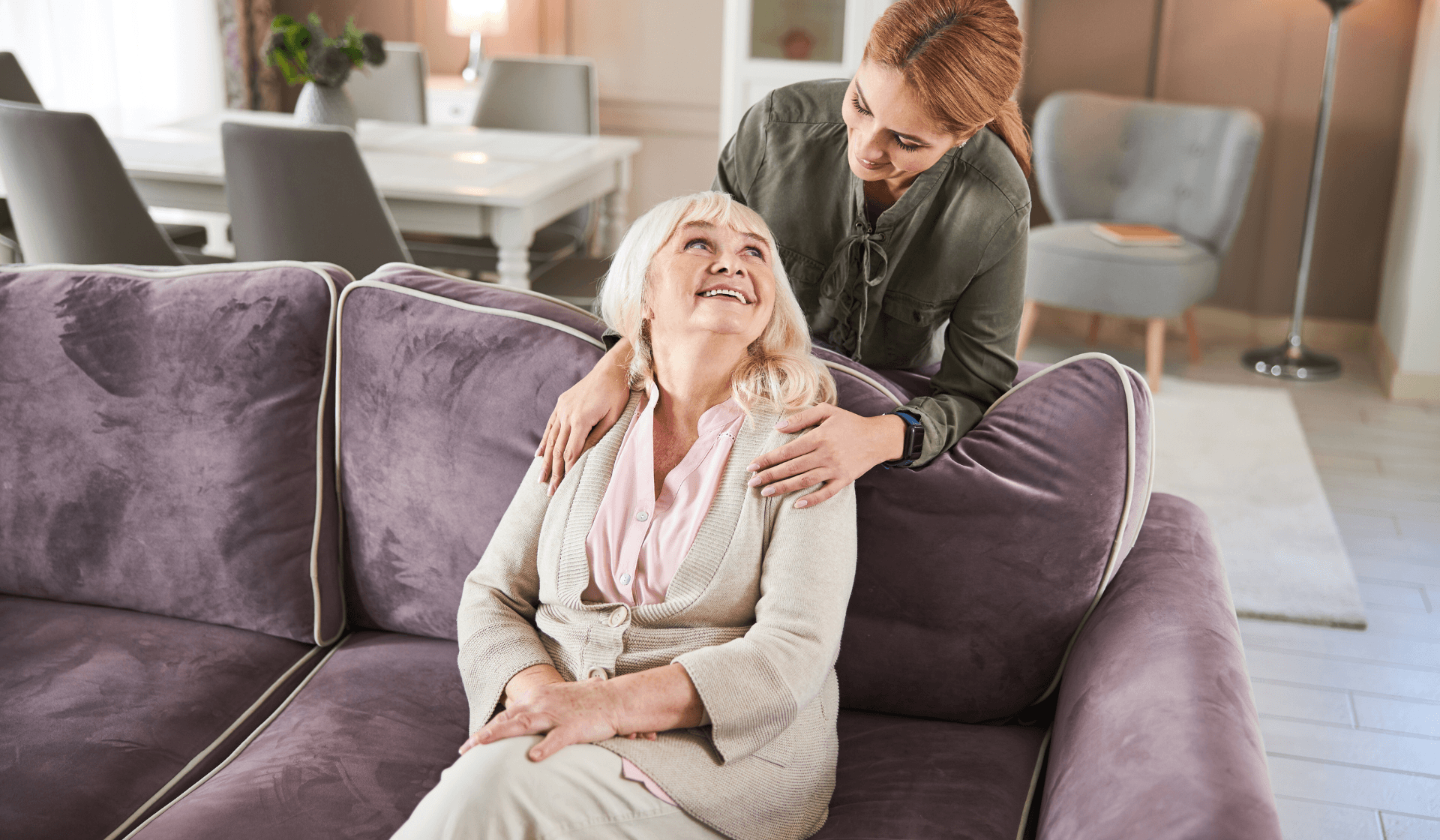 old lady sitting on a couch smiling up at a woman holding her shoulders